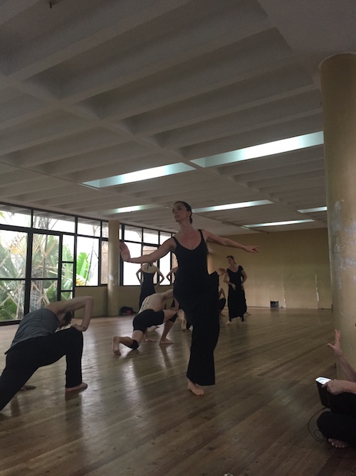 Graham dancers in a Cuban dance studio. A women in a black leotard and pants stands and extends her arm in front of her. Her wrist is flexed.